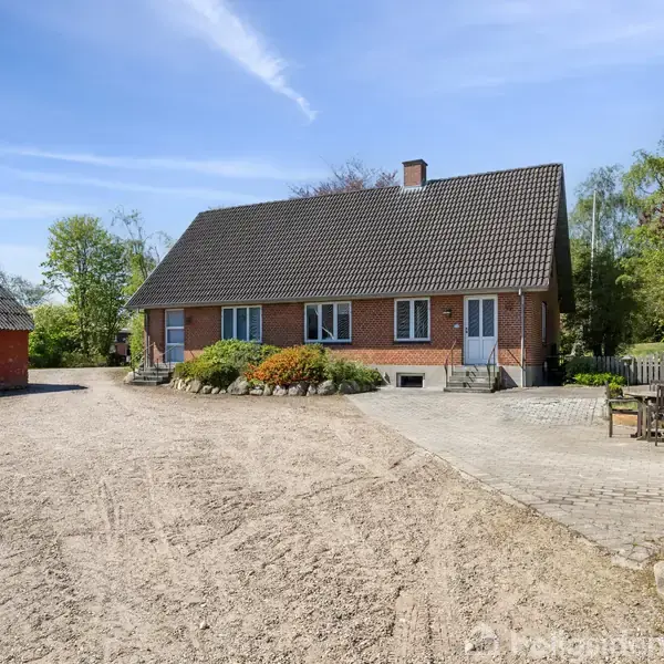 En rød murstensbygning med et sort tegltag står på en gårdsplads omgivet af grus og grønne træer. Til højre ses en bænk og grussti.