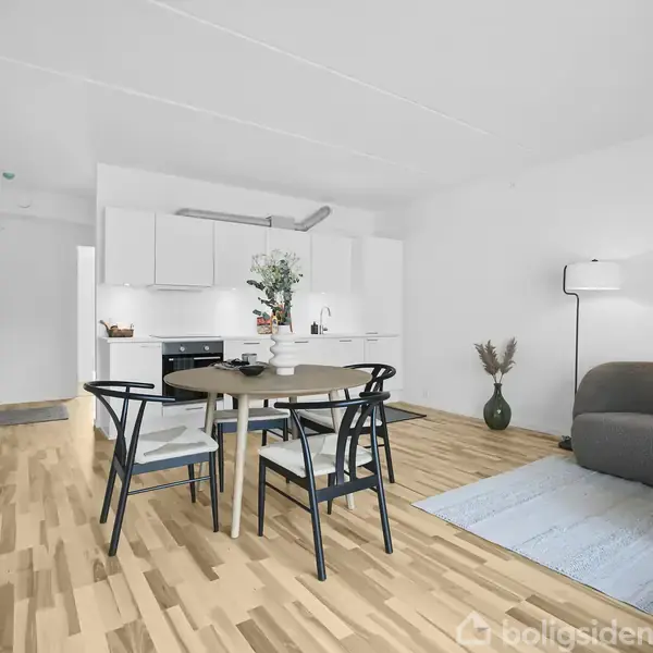 Et moderne køkken-alrum med et rundt spisebord og fire stole i forgrunden. Baggrunden har et åbent køkken med hvide skabe. Til højre er en sofa og gulvlampe på et lyst trægulv.