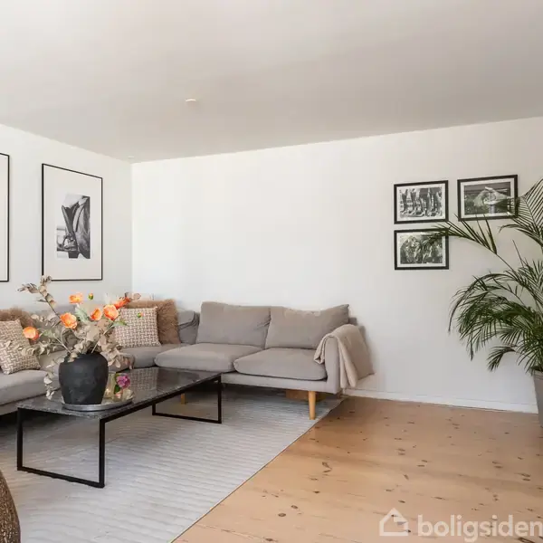 Stue med grå hjørnesofa, to billeder på væggen, og et sofabord med blomster. Plante i vindueskarmen til venstre, lyst trægulv og hvide vægge.