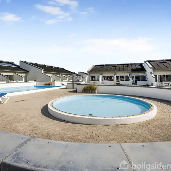 Rund pool med lavt vand står stille i et åbent gårdmiljø omgivet af terrasserede bygninger under en blå himmel. To solsenge står på flisebelægningen ved poolen.