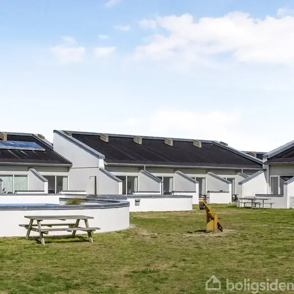 Flere huse med skrånende tage står ved siden af hinanden på en græsplæne. En rund pool og et picnicbord er foran husene under en blå himmel med skyer.