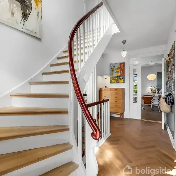 Trappe med trætrin og hvidt gelænder drejer opad til venstre i en lys entré. Vægdekoration og farverige kugler til højre; skænk og rum i baggrunden.