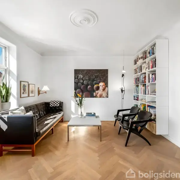 Stue med sort sofa og lave stole omkring et sofabord; vindueskarmen har planter, reolen har bøger. Væggene er dekoreret med kunst, og gulvet er lyst træ.