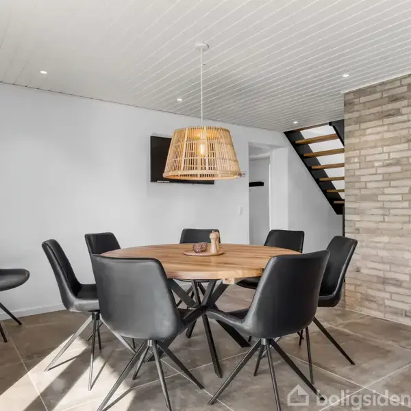 Rundt træbord med seks sorte stole står i et moderne rum. En flettet lampe hænger over bordet. Baggrunden har en murstensvæg og en trappe fører til en anden etage.