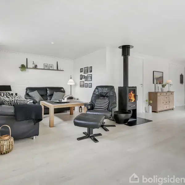 Stue med sort lænestol og pejs, omgivet af sofa, sofabord og vægkunst i et lyst rum med trægulv; til højre ses spiseområde med træmøbler.