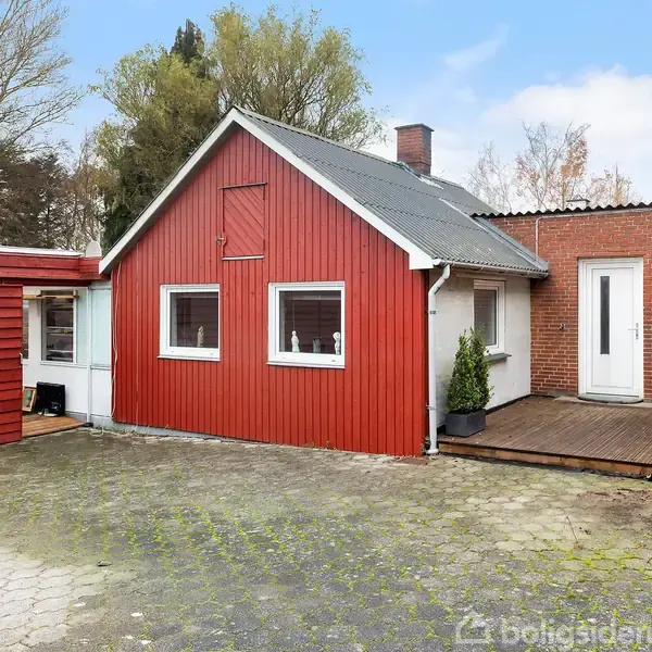 Et rødt træhus med hvide vinduer står stille i en flisebelagt gårdsplads omgivet af træer og en blå himmel.