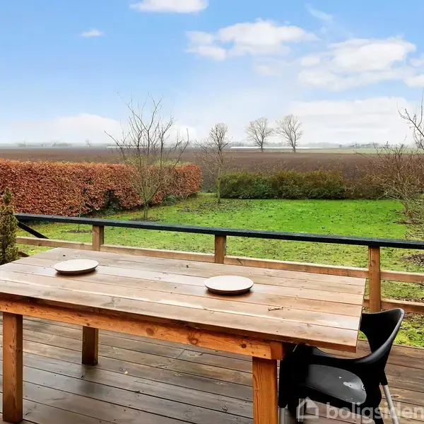 Træbord med to tallerkener står stille på en terrasse med sort stol ved siden af, omgivet af grønne marker og buske under en blå himmel med skyer.