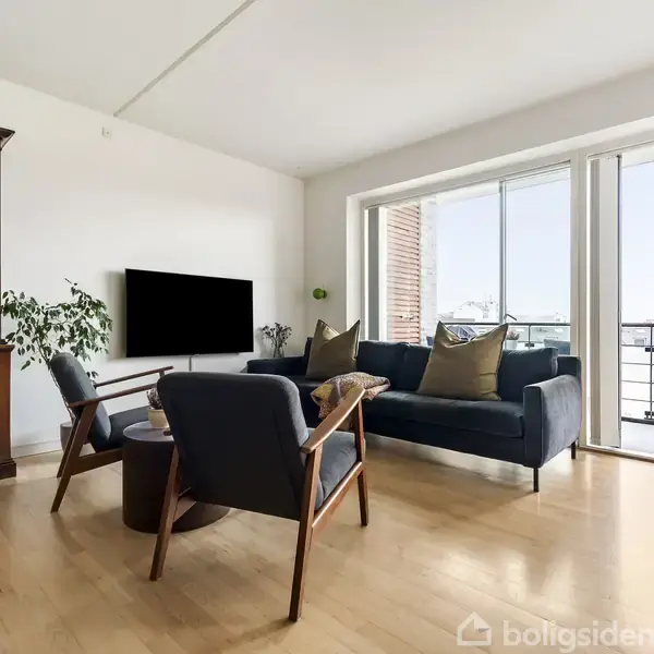 Sofa og stole står i en lys stue med trægulv, placeret foran en fladskærms-TV på væggen. Store vinduer med udsigt, træskab i hjørnet.