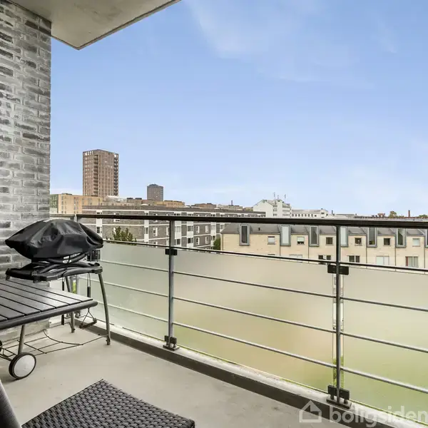 Balkon med møbler og grill står stille i bymæssigt miljø, omgivet af moderne lejlighedsbygninger. En blomst i potte ses på bordet, og en klar blå himmel danner baggrund.