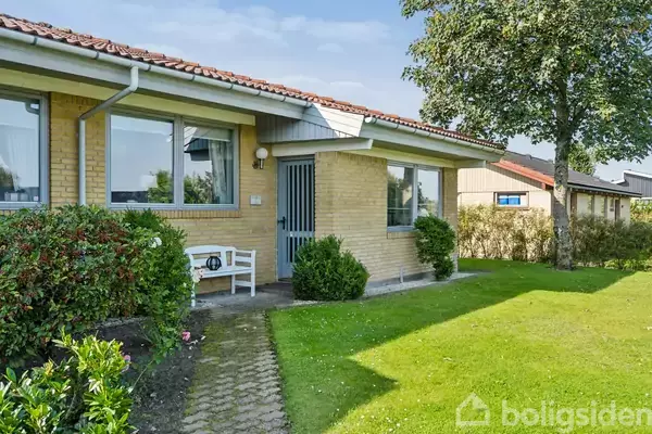 Et gult murstenshus med hvide vinduer og en bænk står i ro. Forhaven består af græsplæne, buske og et træ.