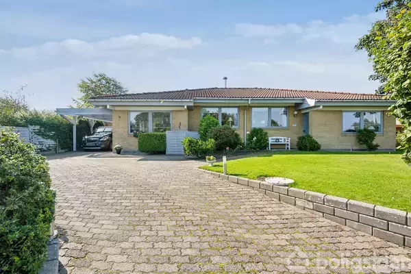 Et enfamiliehus står stabilt med gul murstensfasade, omgivet af en grøn have. Bilen er parkeret under en carport til venstre, og en flisebelagt indkørsel leder fra vej til hus.