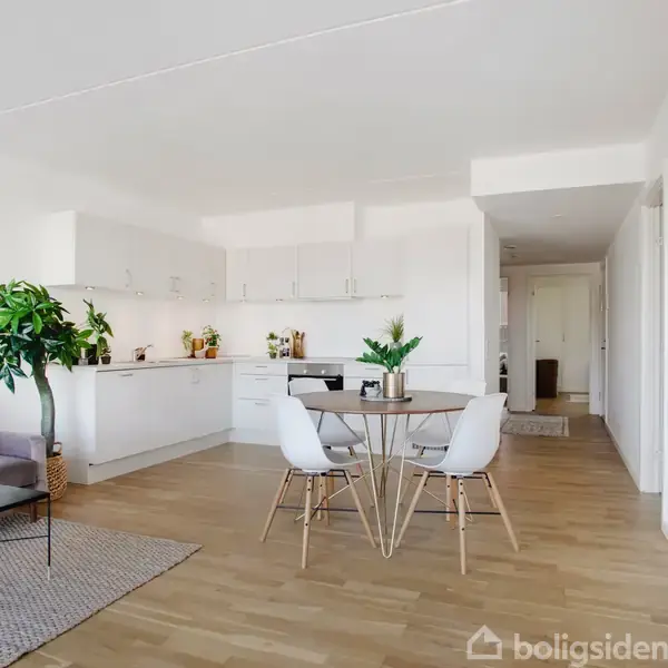 Stue-køkken-alrum med spisebord og stole centralt. Lænestol og bord med planter ved siden. Moderne, hvidt køkken i baggrunden med pynt og køkkenartikler. Mønstret trægulv.