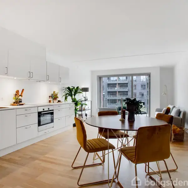 Et moderne køkken-alrum med hvide skabe og træmøbler. Rundt spisebord med fire stole centreret. Stue med sofa ved vindue i baggrunden. Plante dekorationer på bord og i hjørnet.