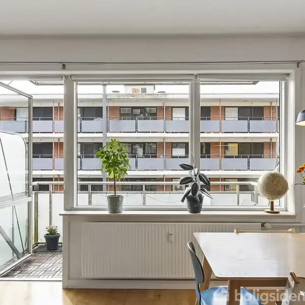 Et spisebord står i et veloplyst rum med udsigt til en altan og en moderne bygning. På bordet er der en vase med blomster. Radiator og planter vises i vindueskarmen.