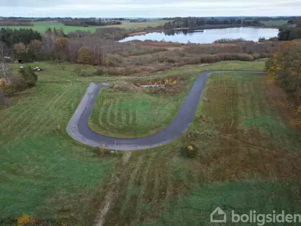 En asfalteret vej former sig som et "U", omgivet af grønne marker og skov. I baggrunden ses en sø og tæt bevoksning.