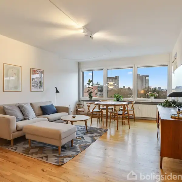 En lys stue med en grå sofa og sofabord placeret på et mønstret tæppe. Til højre står et spisebord med stole foran store vinduer, der giver udsigt til byens bygninger. En trækommode med lampe står i højre side. To plakater hænger på væggen.