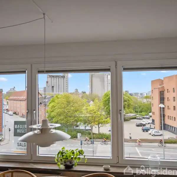 Tre vinduer viser byudsigt med grønne træer, rødbrune bygninger og biler. En lampe hænger indendørs. Tekst på skilt i midten: "MÆGLER HUSET SALG."