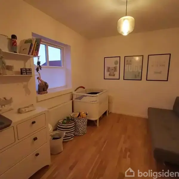 Et børneværelse med en krybbe står langs væggen. Ved siden af er der en grå sofa. Værelset har trægulv, en lysekrone, og opslagstavler på væggen.