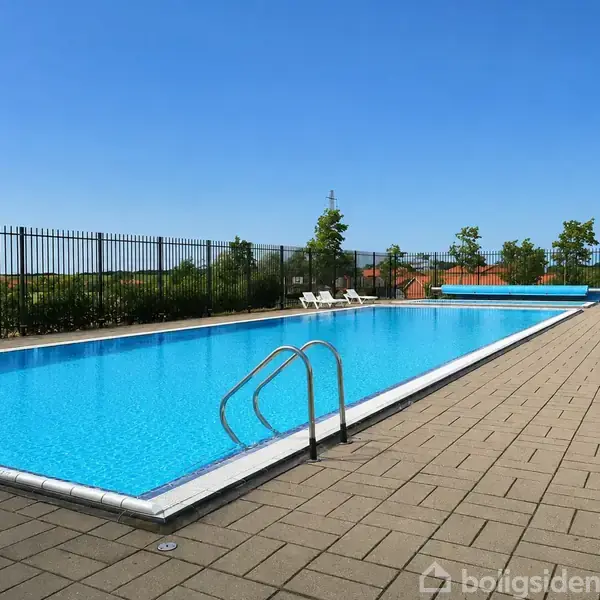 En rektangulær swimmingpool står stille i en udendørs gårdhave med fliser. Baggrunden har en metalhegn, et par liggestole og nogle buske og træer under en klar blå himmel.