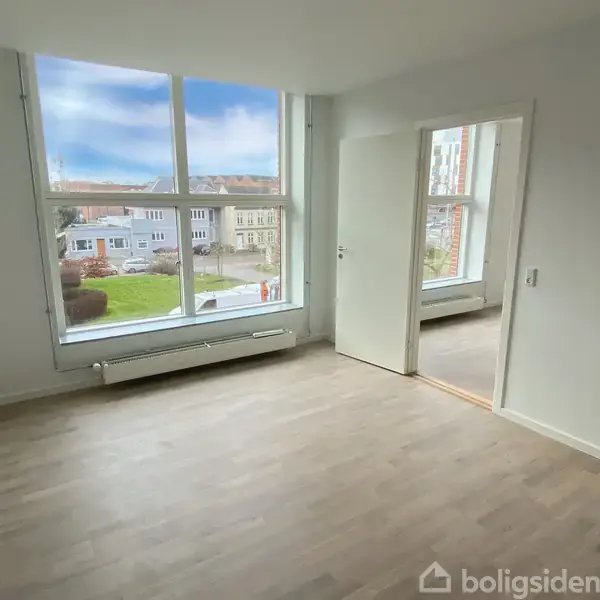 Et lyst rum med trægulv har en stor vinduesvæg. En åben dør leder til endnu et rum. Udenfor ses en udsigt over en grøn have og flere bygninger.