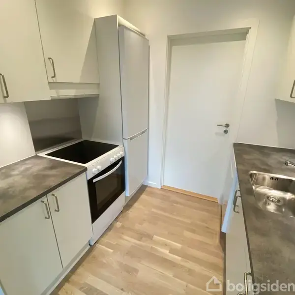 Køkken med moderne, hvide skabe, sort kogeplade, ovn og køleskab. En stålvask er monteret på en grå bordplade, i et rum med lyst trægulv og en hvid dør i baggrunden.