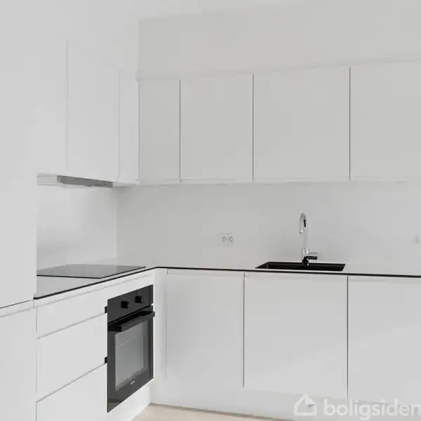 Et moderne køkken med hvide skabe og sort bordplade, indbygget ovn og kogeplade, ved siden af en vask med en stålvandhane, omgivet af hvide vægge og minimal indretning.