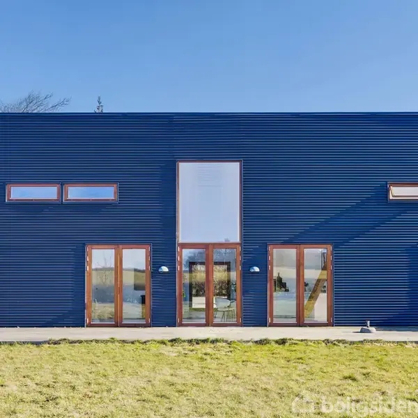 Et moderne, firkantet hus med blå facade og store vinduer står på en grøn græsplæne under en blå himmel. Trævinduesrammer tilføjer varme til designet.