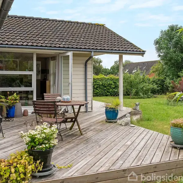 Træterrasse med bord og stole samt planter i potter, beliggende ved en hvidmalet bolig. Haven er grøn med buske og træer i baggrunden på en solrig dag.