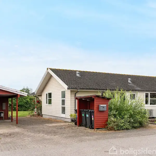 Et enfamiliehus med hvid facade og mørkt tag står ved en have med buske. En carport med rødt træværk står i indkørslen ved siden af huset under en blå himmel.