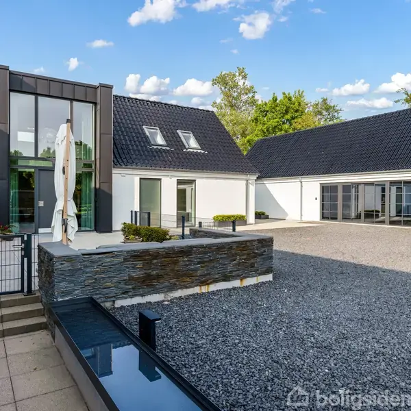 Et moderne hus med sorte tagsten og store vinduer står omgivet af grusbelagt gårdsplads og flisebelagt terrasse. Et vandspejl og en stor parasol pryder indgangen.