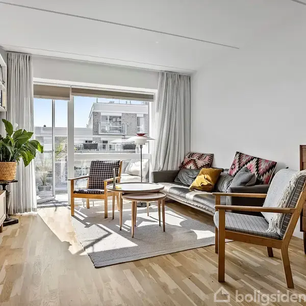 Stue med sofa og stole arrangeret omkring sofaborde, beliggende på et lyst trægulv. Store vinduer slipper lys ind, mens en plante og reol tilføjer dekoration.