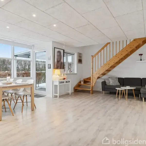 Et lyst stuerum med en trappe af træ, der leder til en overetage. Et spisebord med seks stole står til venstre. Store vinduer giver udsyn til sneklædte buske udenfor.