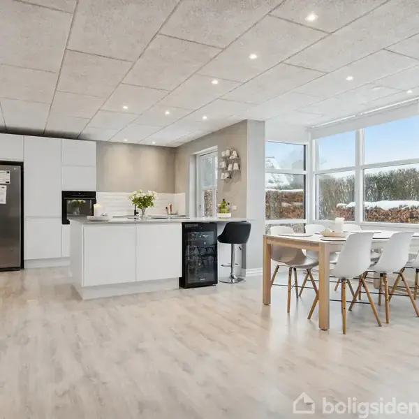 Et moderne køkken med hvidt interiør og spisebord. Stort vindue giver udsigt til sneklædt landskab. Køleskab og ovn er indbygget langs væggen. Lys trægulv fuldender rummet.