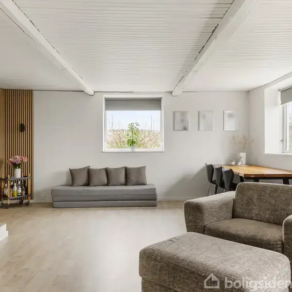 En grå sofa med pudder står ved en væg med kunstmalerier. I rummet er der også en lænestol og et træbord ved vinduet, der skaber en lys, moderne atmosfære.