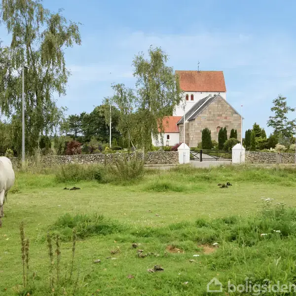 En hvid hest græsser på en grøn mark foran en kirke med rød tegltag, omgivet af træer og stenmur under en blå himmel.