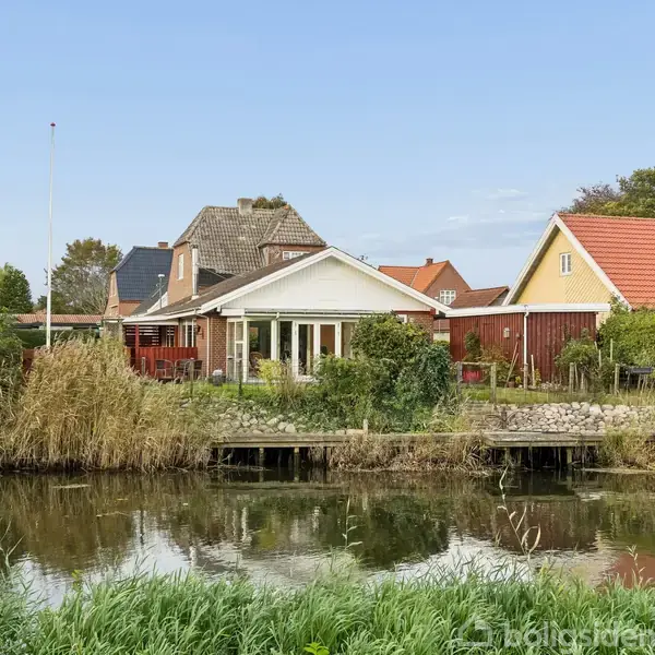 Et hus med en udestue ligger ved en stille sø eller kanal. Omgivet af siv og buske, ses flere huse i baggrunden under en blå himmel.