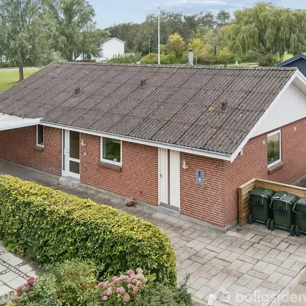 Et rødt murstenshus med skråt tag står stille i et villakvarter. Foran er en flisebelagt indkørsel og en hæk, omgivet af grønne områder og flere huse i baggrunden.