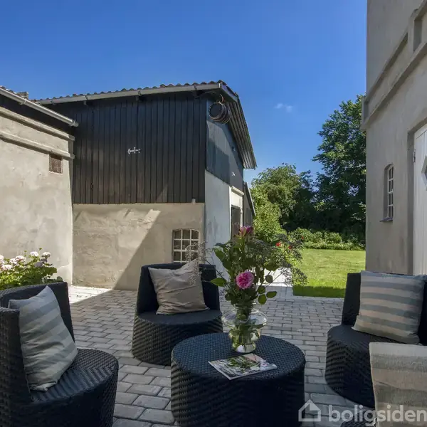 Runde kurvemøbler med puder står på en brolagt terrasse. Et bord med en blomsterdekoration i midten. Et lyst hus og en bygning i træ er i baggrunden.