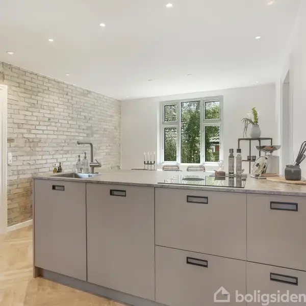 Et moderne køkken med en stor ø med grå skuffer, dekoreret med køkkenredskaber og en plante. I baggrunden er der en murstensvæg, et vindue og lyst trægulv.