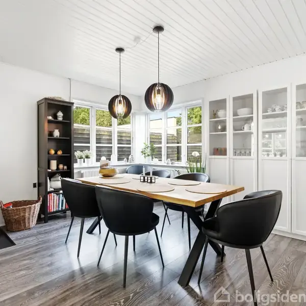 Moderne spisebord med sorte stole omkring; to hængelamper over bordet. Brændeovn i hjørnet; store vinduer med udsigt til have. Reol fyldt med porcelæn. Trægulv og hvide vægge.