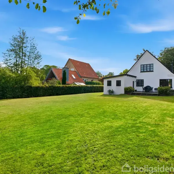 Et hvidt hus står roligt i en stor, velplejet have omkranset af buske. Andre huse med røde tage er synlige i baggrunden under en blå himmel.