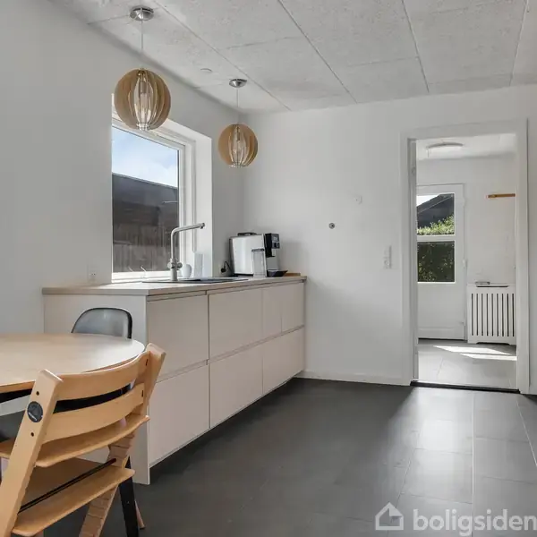 Køkken med lyst interiør har et rundt bord med stole, vask ved vindue og moderne apparater. Lyse vægge og loft samt store vinduer giver rummet et åbent og indbydende miljø.