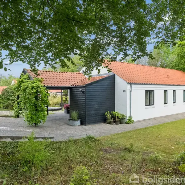 Køkken med træskabe og hvide bordplader. I baggrunden ses en åben reol med tallerkener og en træspisebordsstol. Rødbrune fliser på gulvet og hvidmalet loft. Ingen tekst til transcription.
