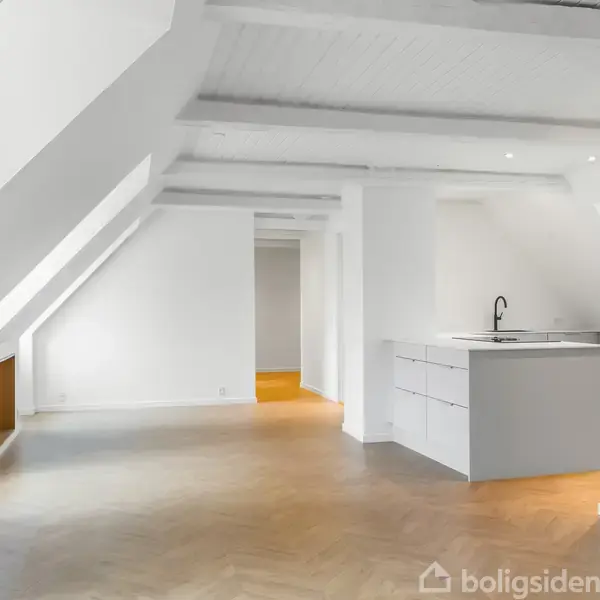Åbent køkken med en ø og indbygget vask, placeret under et skråt loft med synlige bjælker, i en lys, rummelig lejlighed. Hvidmalede vægge og trægulve dominerer rummet.