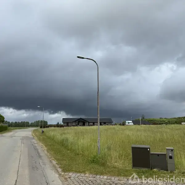 En vejstrækning med en gadelampe strækker sig gennem en mark. I baggrunden ses et hus og en bil under mørke skyer, hvilket antyder et muligt regnvejr.