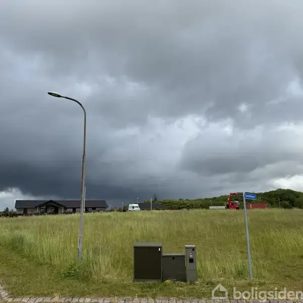 En gadelampe står stille ved en vej i et åbent, grønt område med en mørk, skyet himmel. Der er et hus og køretøjer i baggrunden. Skiltet viser "Rolighedsvej".
