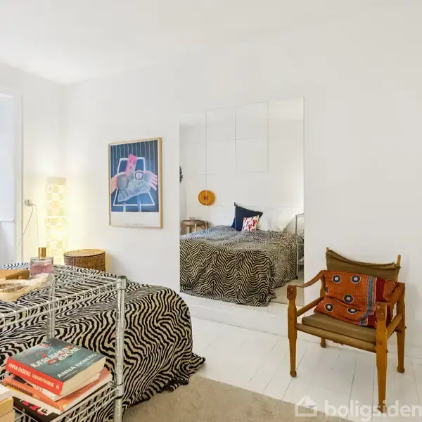 En stue med en grøn sofa, sofabord og en lyserød sækkestol. Et spisebord står ved vinduet med udsigt til boligblokke. Kunst på væggene og planter i hjørnet.