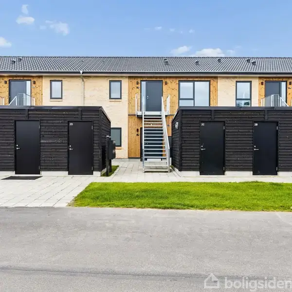 Rækkehuskompleks med trapper førende til øvre etage, omgivet af små sorte skure i forgrunden på en flisebelagt gangsti, med græsplæner og blå himmel i baggrunden.