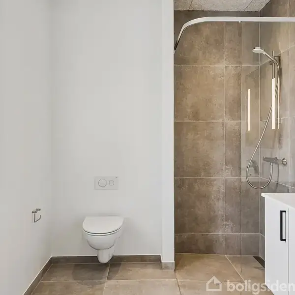 Et lyst badeværelse indeholder en væghængt toilet, glasafskærmet bruser i hjørnet og en moderne vask med skab. Rummet har store, lyse fliser og et vindue til venstre.