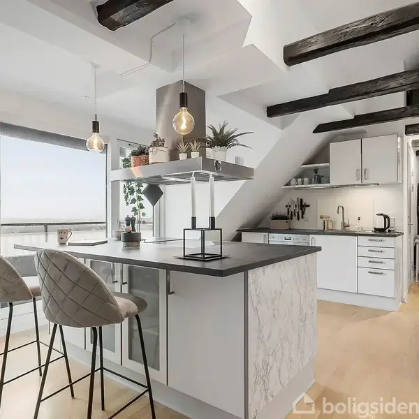 Et moderne køkken med en køkkenø, hvor to høje stole står. Køkkenet har hvide skabe, sorte bjælker i loftet og en stor altandør, der giver udsigt over landskabet.
